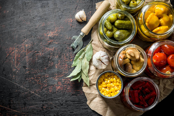 Pickled vegetables in jars on the paper with the Bay leaves and the cloves of garlic. On dark rustic background - Fotoğraf, Görsel