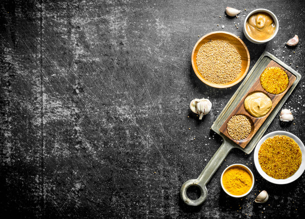 Different types of mustard in plates and on a stand with garlic slices. On dark rustic background - Fotoğraf, Görsel