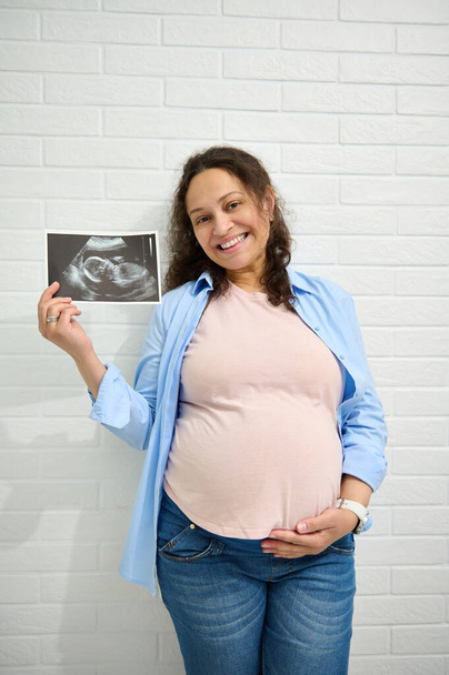 Feliz afro-americano positivo mulher grávida adulta, acariciando sua barriga grande, sorrindo e mostrando à câmera o ultra-som de seu bebê recém-nascido, isolado sobre fundo de parede de tijolo branco. Gravidez - Foto, Imagem