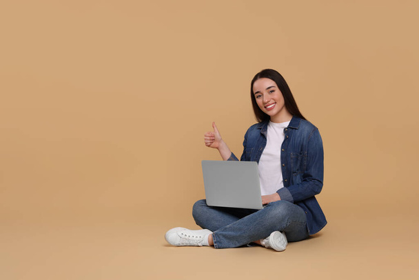 Smiling young woman with laptop showing thumbs up on beige background, space for text - Photo, Image