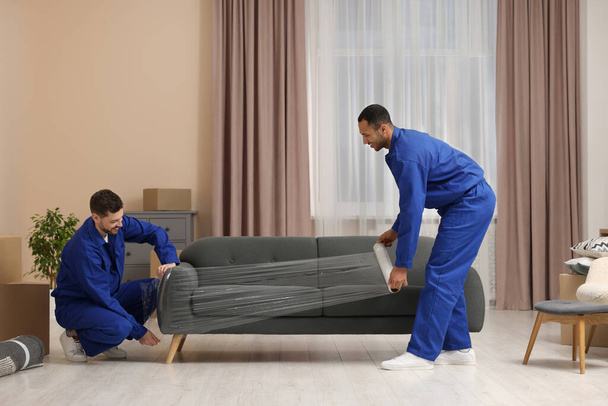 Male movers with stretch film wrapping sofa in new house - Zdjęcie, obraz