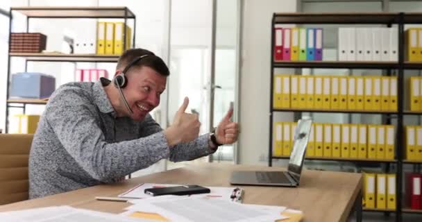 Een lachende zakenman in een koptelefoon kijkend naar een laptop scherm, kijkend naar een goede webinar die duimen opsteekt. Mood swings consulting client of het leren van talen op afstand tonen gebaar fak deelnemen aan - Video