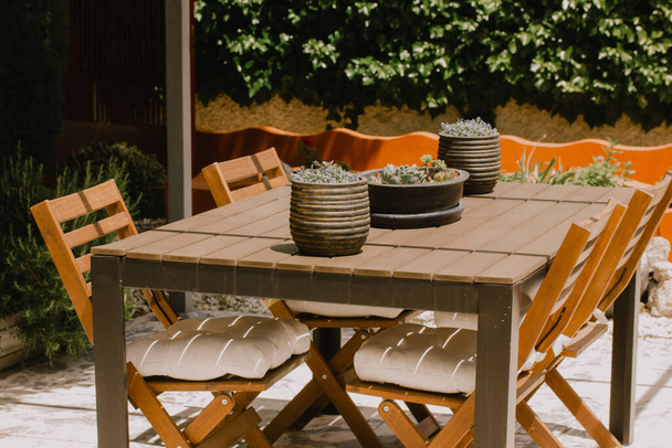 Gartenmöbel im Freien. Holztisch, Stühle im Hinterhof. Dekorative Blumentöpfe stehen auf dem Tisch. Terrassenhof tagsüber. Lounge-Platz zum Entspannen im Freien an sonnigen Tagen. Stühle aus braunem Holz - Foto, Bild