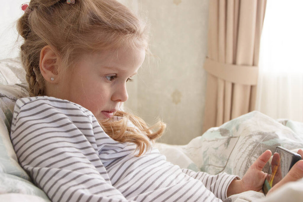 Egy aranyos kölyök fekszik az ágyban okostelefonnal a kezében. Az otthoni oktatás fogalma - Fotó, kép