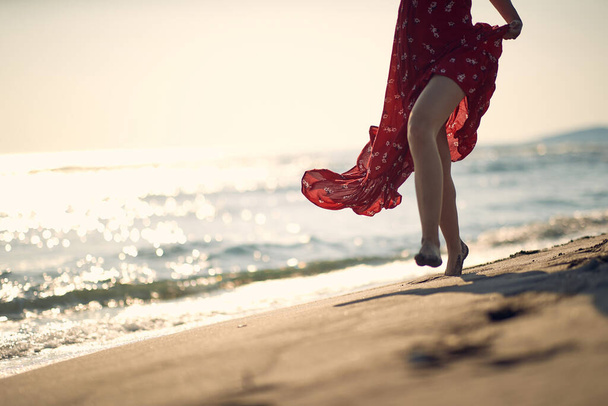 Young woman walking on the shore of beach. Woman in red dress, focus on legs. Fashion, summertime, lifestyle concept. - Фото, зображення