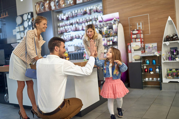 Lovely family of young couple and toddler girl in a home decor shop together, faher high fiving his daughter, family feeling joyful. Shopping, togetherness, lifestyle concept. - Фото, изображение