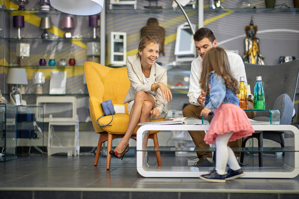 Lovely young couple with their toddler daughter in lamp shop choosing a lamp, looking through a catalogue together. Modern home decor shop. Lifestyle concept. - Valokuva, kuva
