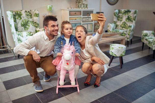 Happy family in a furniture shop, taking a selfie, buying a rocking horse unicorn for toddler daughter. Family, lifestyle, shopping concept. - Foto, imagen