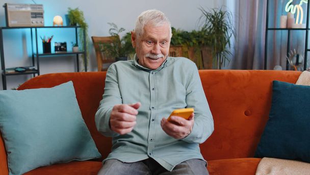 Happy vanhempi isoisä mies käyttää mobiili älypuhelin kirjoittamalla selailu sanoa Vau kyllä selville suuri voitto hyviä uutisia juhlia lotossa jättipotti, voitto ele. Vanhempi kaveri kotona huoneessa istuu sohvalla - Valokuva, kuva