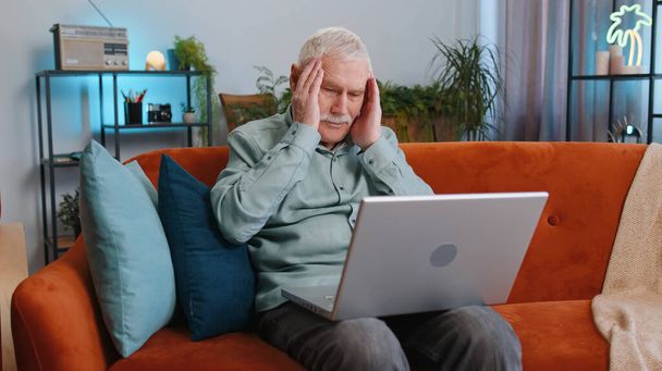 Il nonno stanco l'uomo più anziano usa il computer portatile, soffrendo di tensione di problema di mal di testa ed emicrania, lo stress a casa. Il ragazzo anziano lavora sul taccuino, invia messaggi, fa acquisti online, guarda film - Foto, immagini