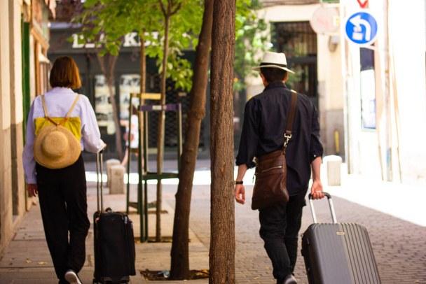 Μαδρίτη, Ισπανία 1 Ιουνίου 2022 Τουρίστες με βαλίτσες και καπέλα περπατώντας σε ένα δρόμο της πόλης πίσω όψη, απαλή εστίαση. Ταξιδεύοντας στην Ευρώπη σε μια καλοκαιρινή περίοδο. Τουρισμός, ταξιδιωτική έννοια Αστικός τρόπος ζωής - Φωτογραφία, εικόνα
