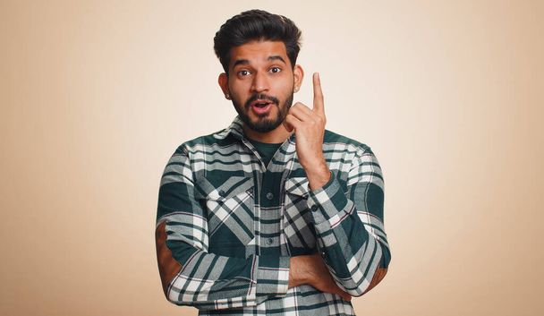 Eureka. Inspired young indian man pointing finger up with open mouth, having good idea, plan, startup, showing inspiration motivation gesture, problem solution. Bearded hindu guy on beige background - Photo, image