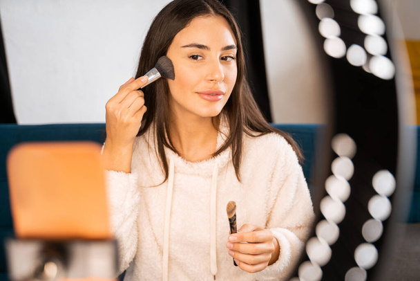Brunette elegant woman blogger sitting in front of the phone and powders her face. She looks into the camera with a smile while recording a video - Photo, Image