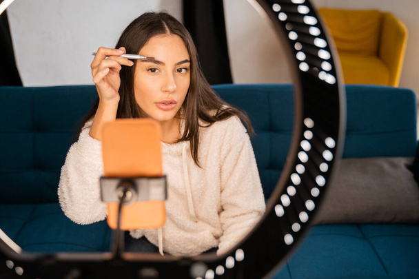 Focused young female blogger in casual clothes sitting on sofa and recording makeup tutorial for social media on smartphone with microphone - Foto, afbeelding
