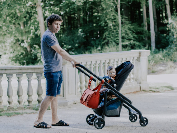 Caucasian young and handsome dad rolls his little daughter sitting in a stroller and looking to the side with a curious look in a public park, close-up side view. Fatherhood concept, fathers, family - Photo, image