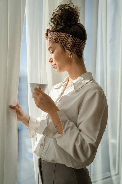 One woman caucasian female standing at he window at home or hotel room in bright day holding a cup of coffee enjoy while taking a brake daily morning routine real people copy space wear white shirt - Photo, Image