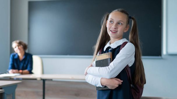 Caucasian girl and female teacher in the classroom. The schoolgirl is holding a textbook - 写真・画像