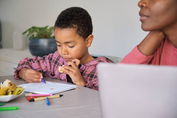 Afrikaanse kleuter jongen met een snack tijdens het schilderen of het doen van het huiswerk thuis, terwijl zijn moeder werkt in een laptop. - Foto, afbeelding