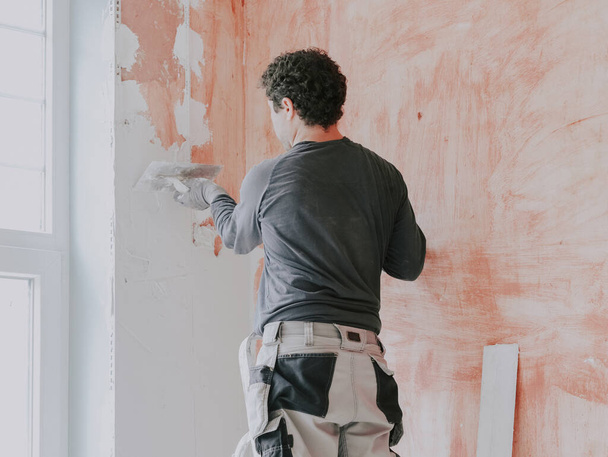 Un joven caucásico con una camiseta gris con mangas, pantalones de trabajo y pelo castaño rizado aplica masilla fresca con una pequeña espátula en la pared cerca de la ventana, de pie sobre una escalera, vista lateral de cerca - Foto, Imagen