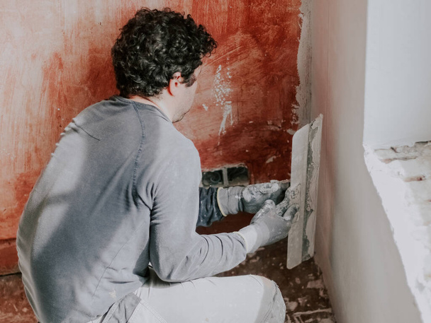 A young caucasian guy in a gray t-shirt with sleeves and curly brown hair checks for evenness a fresh putty on the wall under the window holding a large spatula in his hands, close-up side view. The - Fotografie, Obrázek