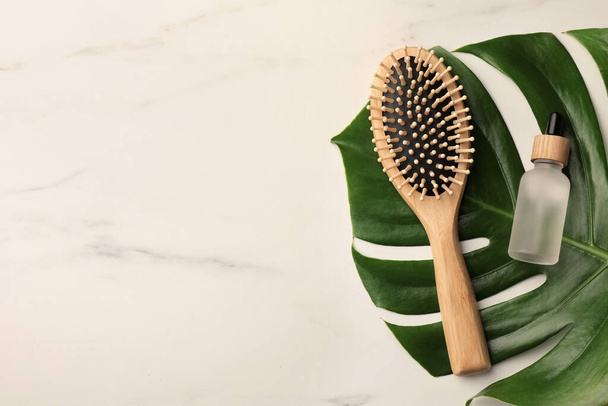 Wooden hair brush, bottle of essential oil and monstera leaf on white marble table, top view. Space for text - Foto, Imagem