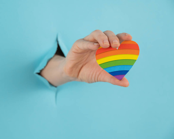 A hand with a rainbow-colored heart sticking out of a hole in a blue cardboard background - 写真・画像