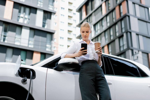 Focus businessman using phone, leaning on electric vehicle, holding coffee with blurred city residential condo buildings in background as progressive lifestyle by renewable and sustainable EV car. - Фото, изображение