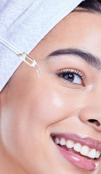 Studio Portrait of a beautiful mixed race woman applying a soothing face serum to her radiant smooth face while wearing a towel on her head. Hispanic woman with flawless skin using essential oil. - Fotografie, Obrázek