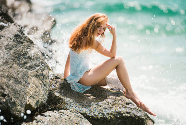 Side view of serene woman sitting on sandy rocky beach against blue waves. Attractive young woman wearing sexy summer dress on a beach - 写真・画像