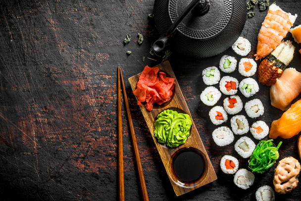 Different types of Japanese sushi, rolls and maki with sauce, wasabi and green tea in a teapot. On dark rustic background - Photo, image