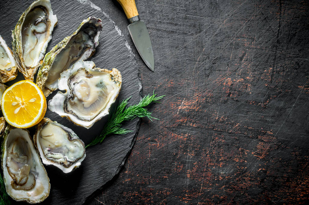 Raw oysters on a round stone Board with lemon. On dark rustic background - Zdjęcie, obraz