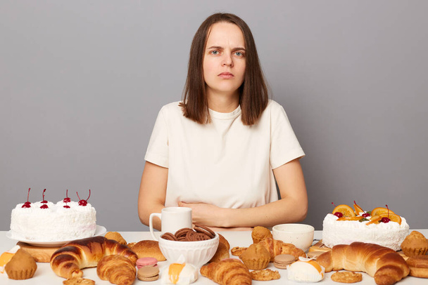 Sad sick woman wearing white T-shirt isolated over gray background sitting at festive table among pastry being overeating feeling pain in belly. - Фото, зображення