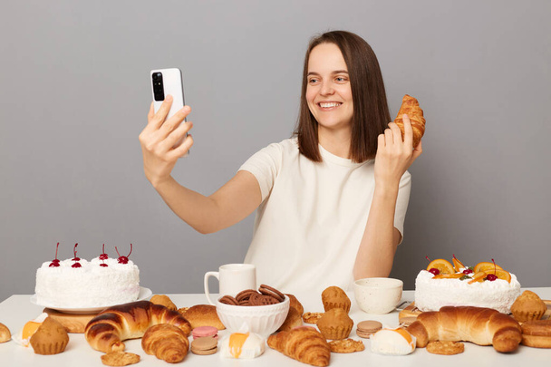 Smiling cheerful joyful woman wearing white T-shirt isolated over gray background sitting at festive table among sugary desserts having video call holding croissant. - Photo, Image
