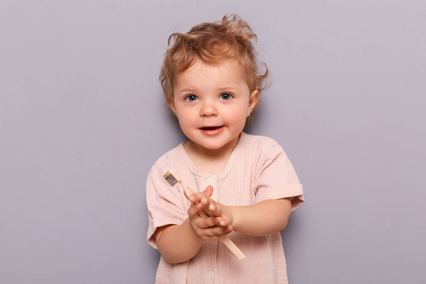 Charming little girl holding toothbrush isolated on gray background looking at camera holding toothbrush trying to brush teeth by herself. - Photo, image