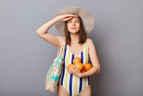 Ernste Frau im Badeanzug isoliert auf grauem Hintergrund stehend mit Orangen hält die Hand in der Nähe der Stirn und sieht weit aus, als warte sie auf ihren Tropenurlaub. - Foto, Bild