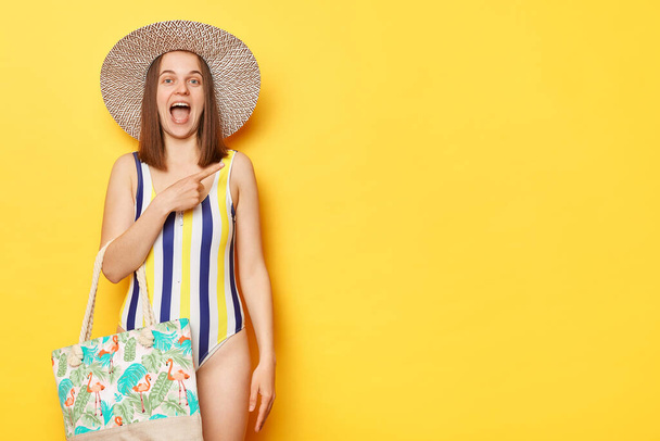 Excited amazed woman wearing swimsuit pointing at advertisement area screaming with excitement astonished promotion isolated on yellow background. - Photo, image