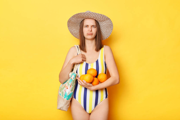 黄色の背景に隔離された水着姿の失意の女性がカメラを見て果実を保持します。. - 写真・画像