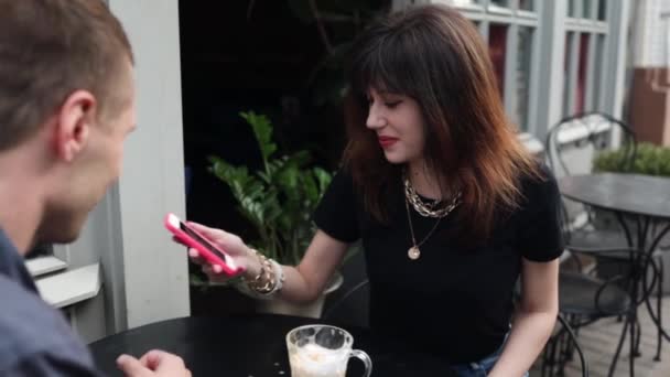 Mujer joven sentada con su novio en la mesa de la cafetería, comiendo y discutiendo algo en el teléfono inteligente - Imágenes, Vídeo