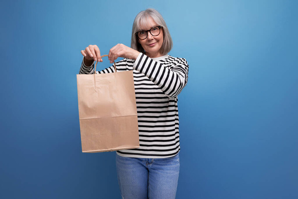コピースペースとの明るい背景にエコフレンドリーなショッピングバッグのモックアップを保持する灰色の髪を持つ愛らしい50代の成熟したビジネス女性. - 写真・画像