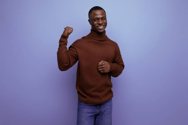 умный темнокожий 25-летний американец с короткой стрижкой в коричневом свитере показывает свою руку на фоне студии с копировальным пространством. - Фото, изображение