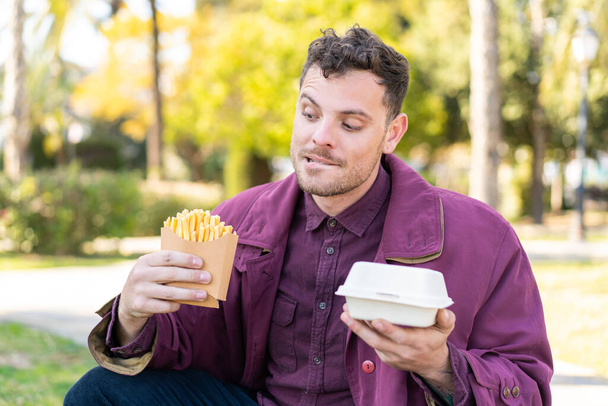 Young caucasian man at outdoors taking fried chips and takeaway box food and thinking - Photo, Image