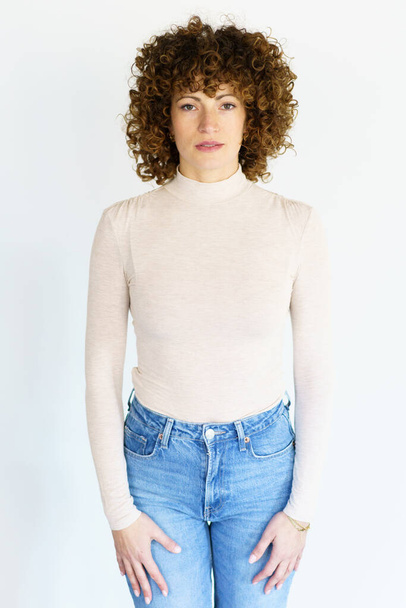 Adult female in beige turtleneck and jeans with brown curly hair looking at camera while standing still with arms hanging straight on sides - Foto, imagen