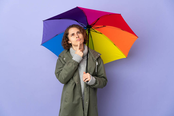 English woman holding an umbrella isolated on purple background having doubts while looking up - Foto, afbeelding