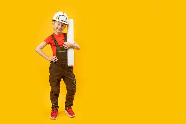 Rapaz de capacete protector, macacão e com folha de papel nas mãos. Criança como construtora ou arquiteta. Espaço de cópia, mockup. - Foto, Imagem
