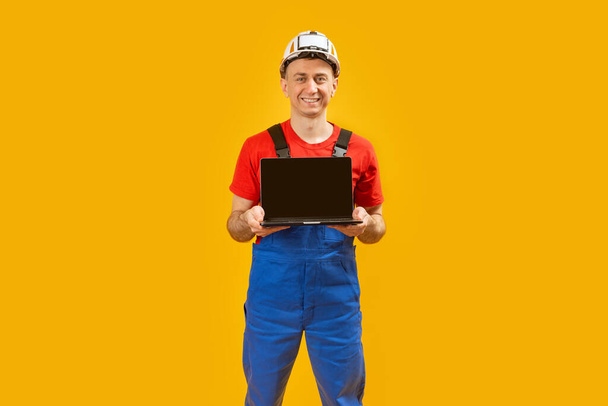 Lächelnder Mann mittleren Alters, Ingenieur im Schutzhelm, Laptop mit leerem Bildschirm. Gelber Studiohintergrund. Kopierraum, Attrappe - Foto, Bild
