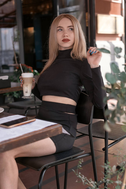 Stylish young woman in tight black clothes sits at table in cafe and holds latte or cappuccino coffee. Vertical frame - Photo, image