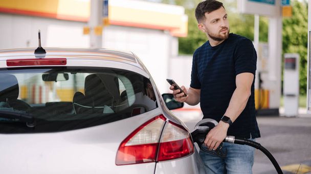 Handsome bearded man refueling car and looking on the scoreboard while standing on self service gas station. High quality photo - Photo, Image