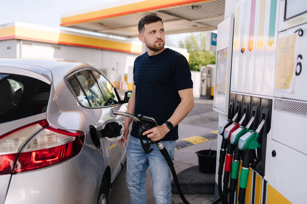 Yakışıklı, sakallı bir adam arabaya yakıt dolduruyor ve benzin istasyonunda dikilirken skorborda bakıyor. Yüksek kalite fotoğraf - Fotoğraf, Görsel