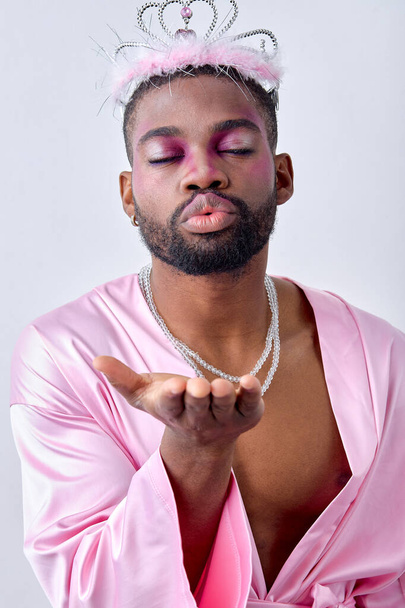 Lovely black american guy in pink shirt and crown flowing a kiss at camera with eyes closed, isolated over white studio background. young man is half-naked, kissing. feminine gay with make-up on face - Foto, imagen