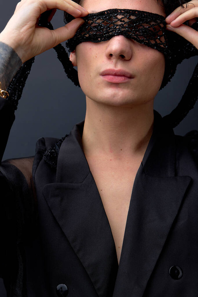 Close-up portrait of young sexy womanlike male in black suit with eye mask, isolated on black studio background. Passionate man is looking fashionable, stylish. fashion, beauty, people concept - Fotoğraf, Görsel
